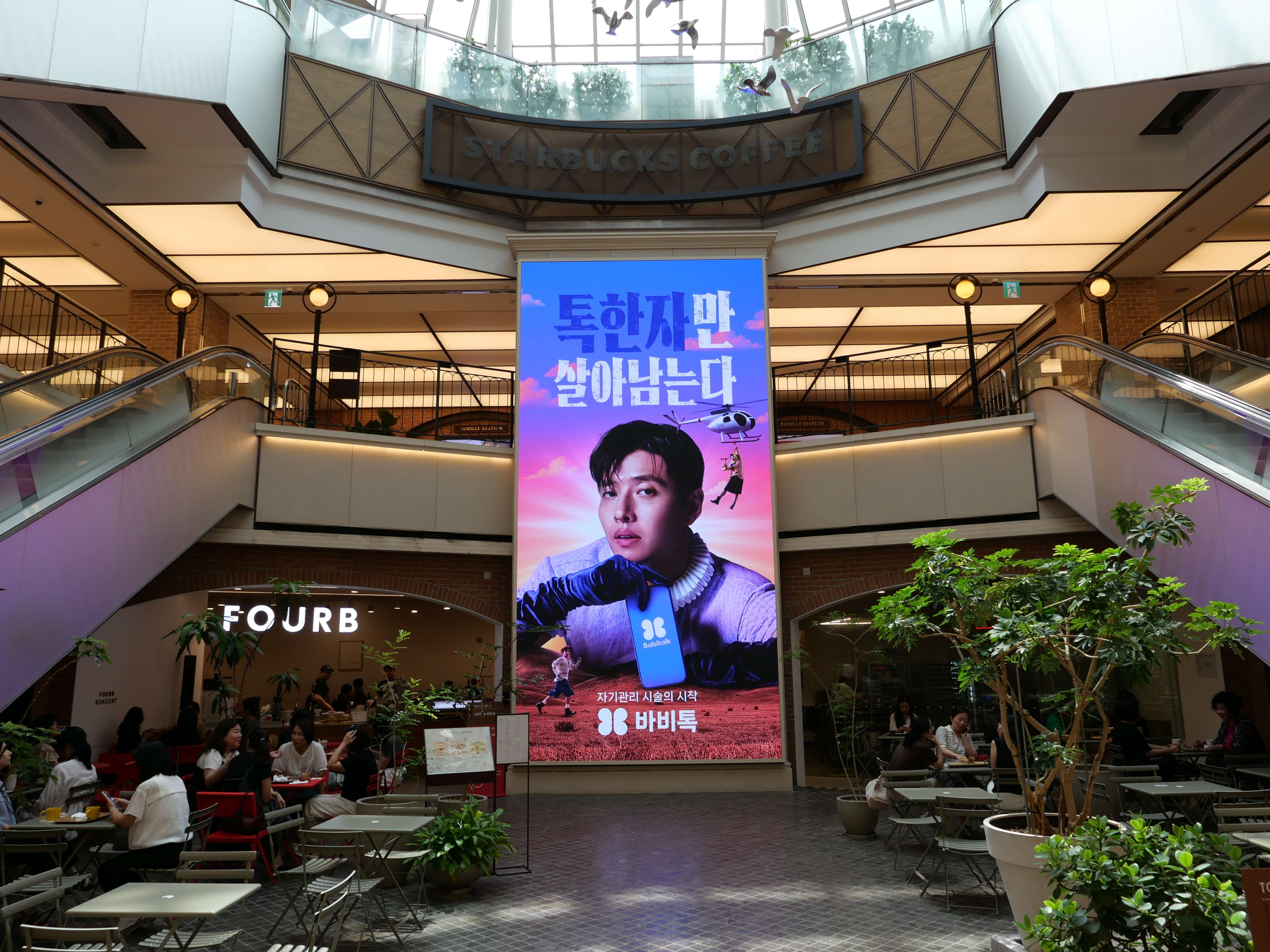 Large vertical BABITALK digital screen rising through the open atrium between escalators at Famille Station, Seoul