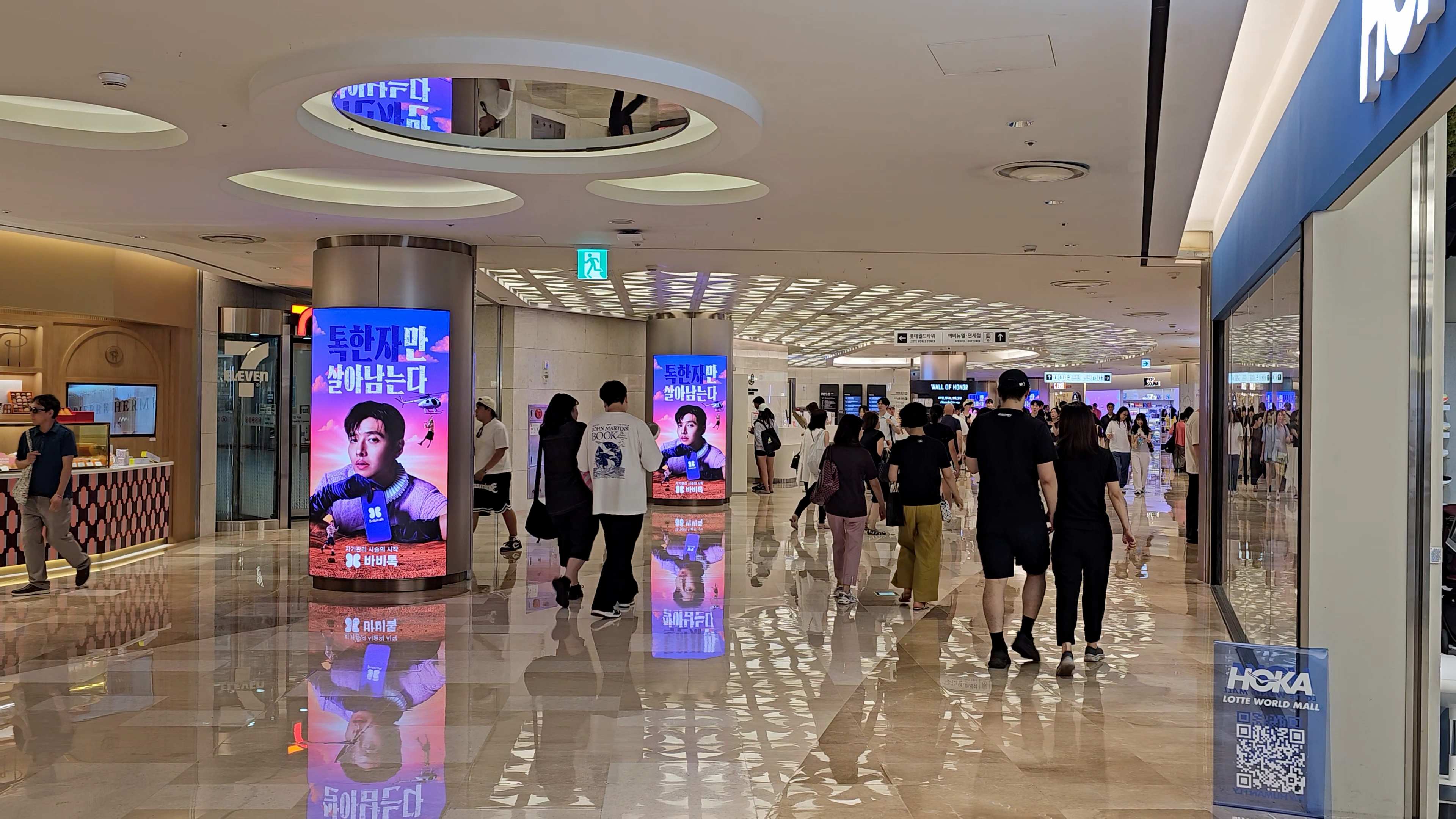 Two curved BABITALK digital columns reflected on the marble floor of a busy Lotte World Mall corridor, Jamsil