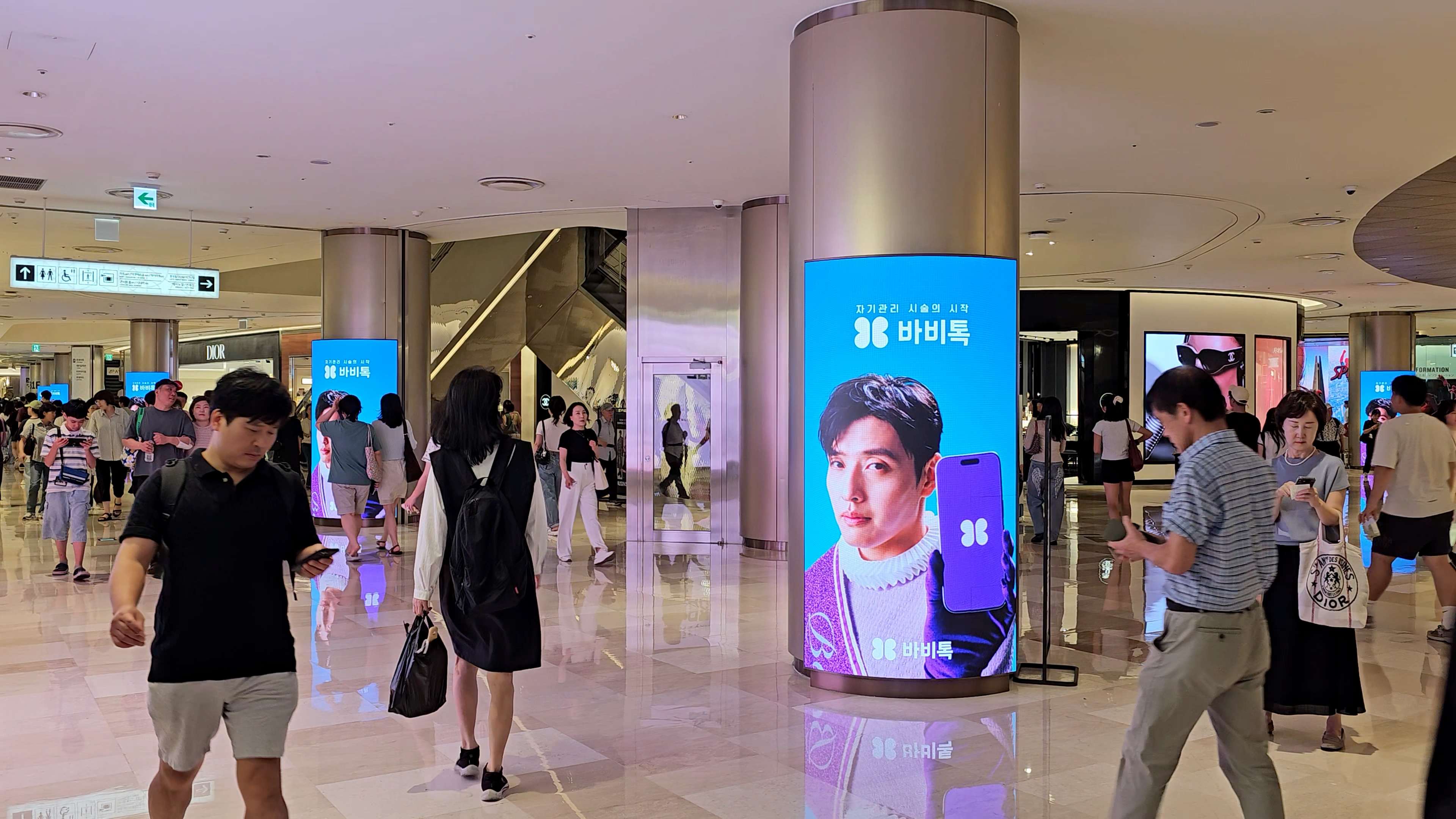 BABITALK creative on a curved digital column among luxury storefronts and passing shoppers, Lotte World Mall, Jamsil