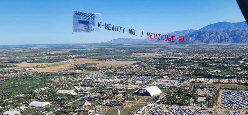 "K-BEAUTY NO. 1 MEDICUBE" aerial banner flying over the Coachella Valley festival grounds and mountains