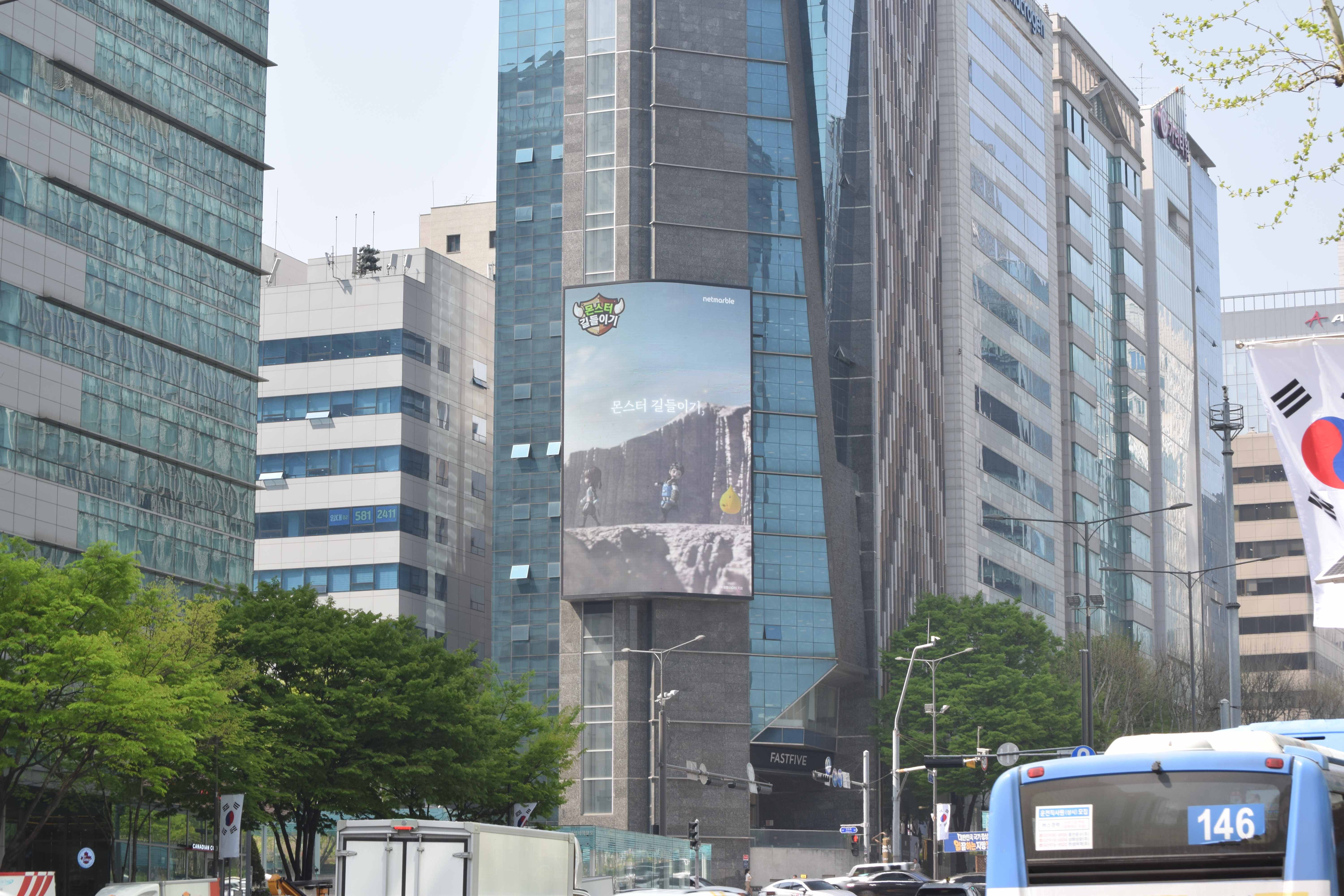 Monster Taming creative on a curved screen mounted on a dark granite tower, with trees and a city bus below, Seoul