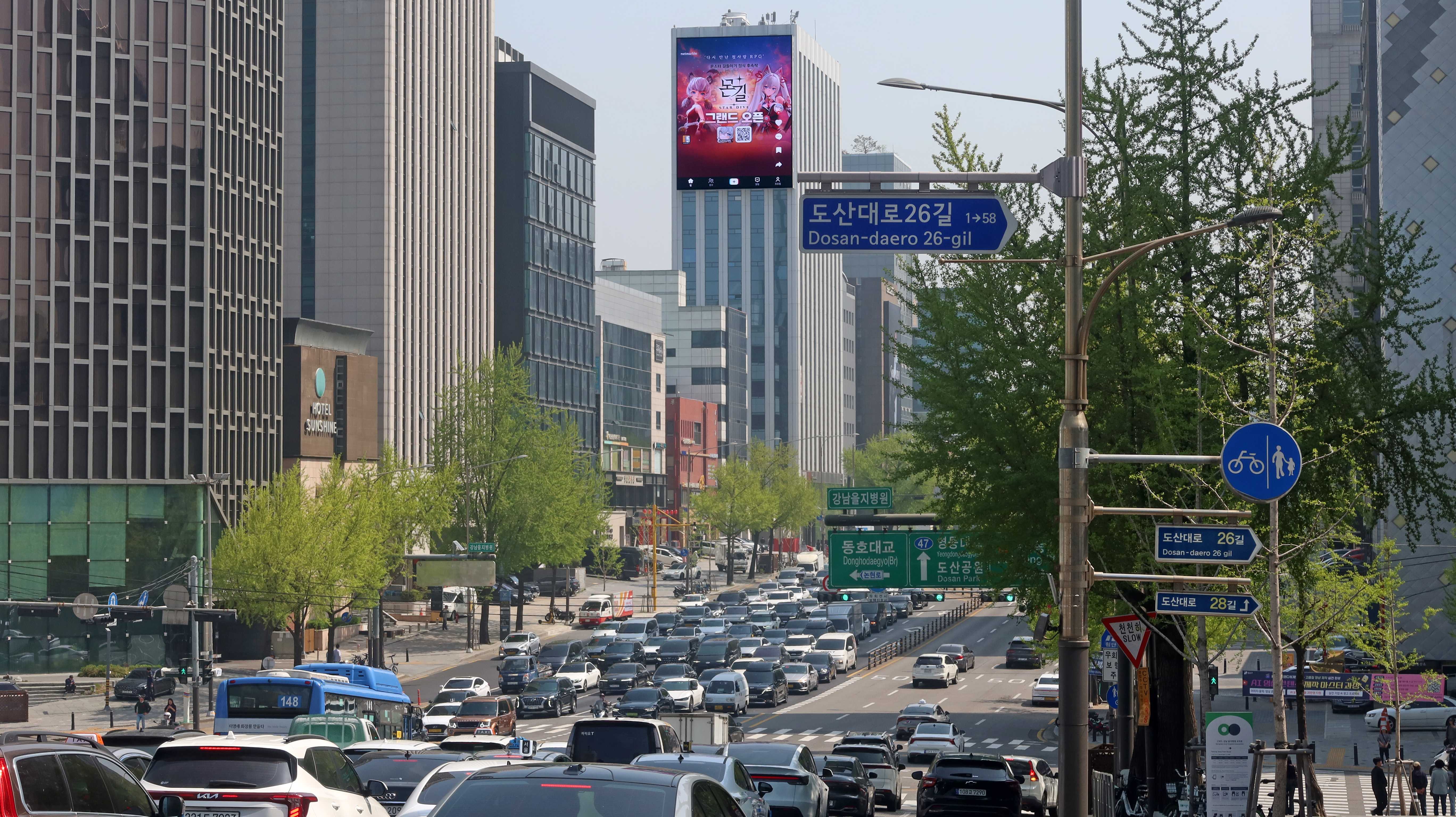 Traffic on Dosan-daero leading toward a tall vertical screen running MONGIL: STAR DIVE grand open creative, Seoul