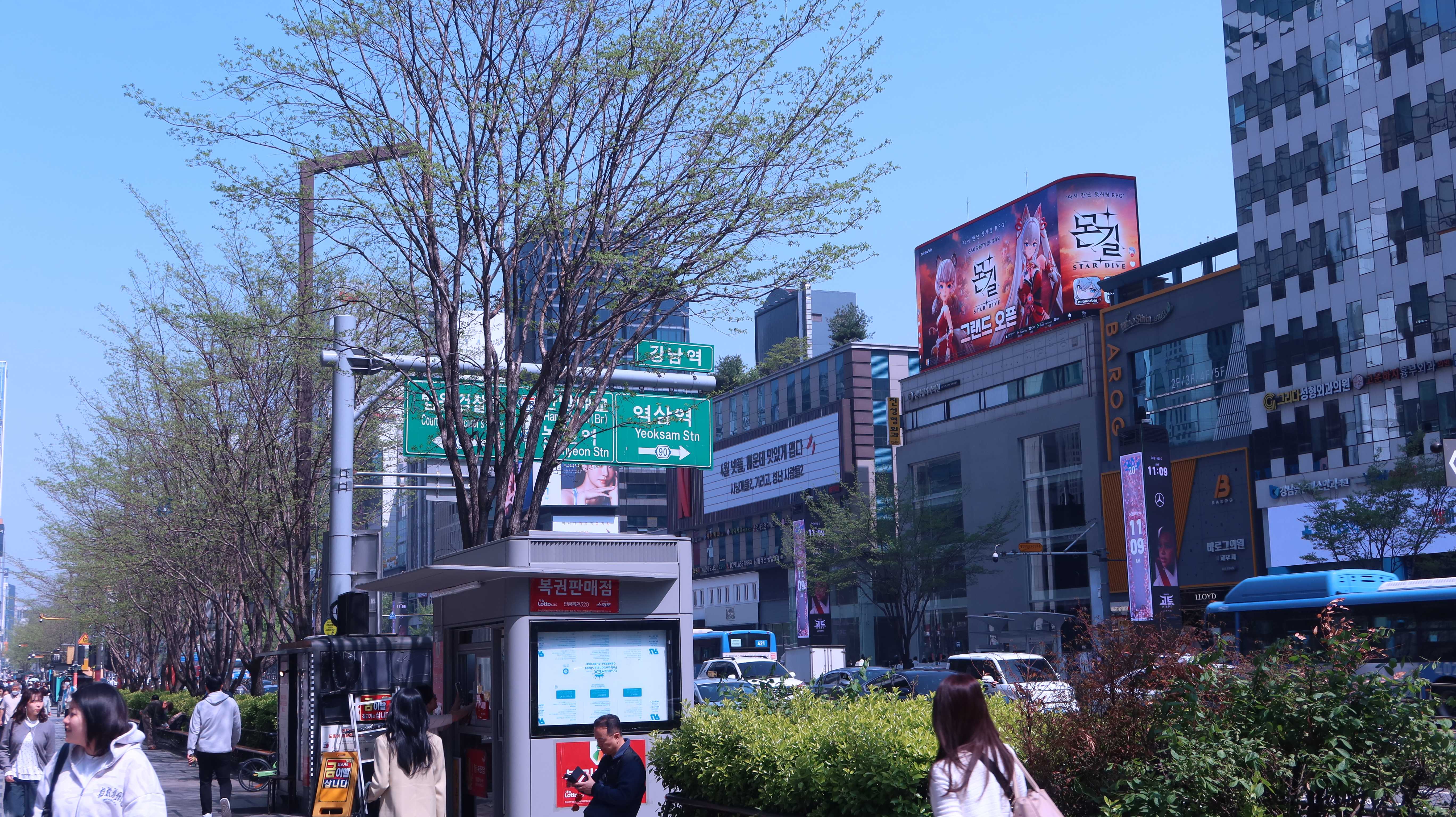 Gangnam Station street with trees, road signs and pedestrians beneath the curved MONGIL: STAR DIVE grand open screen, Seoul