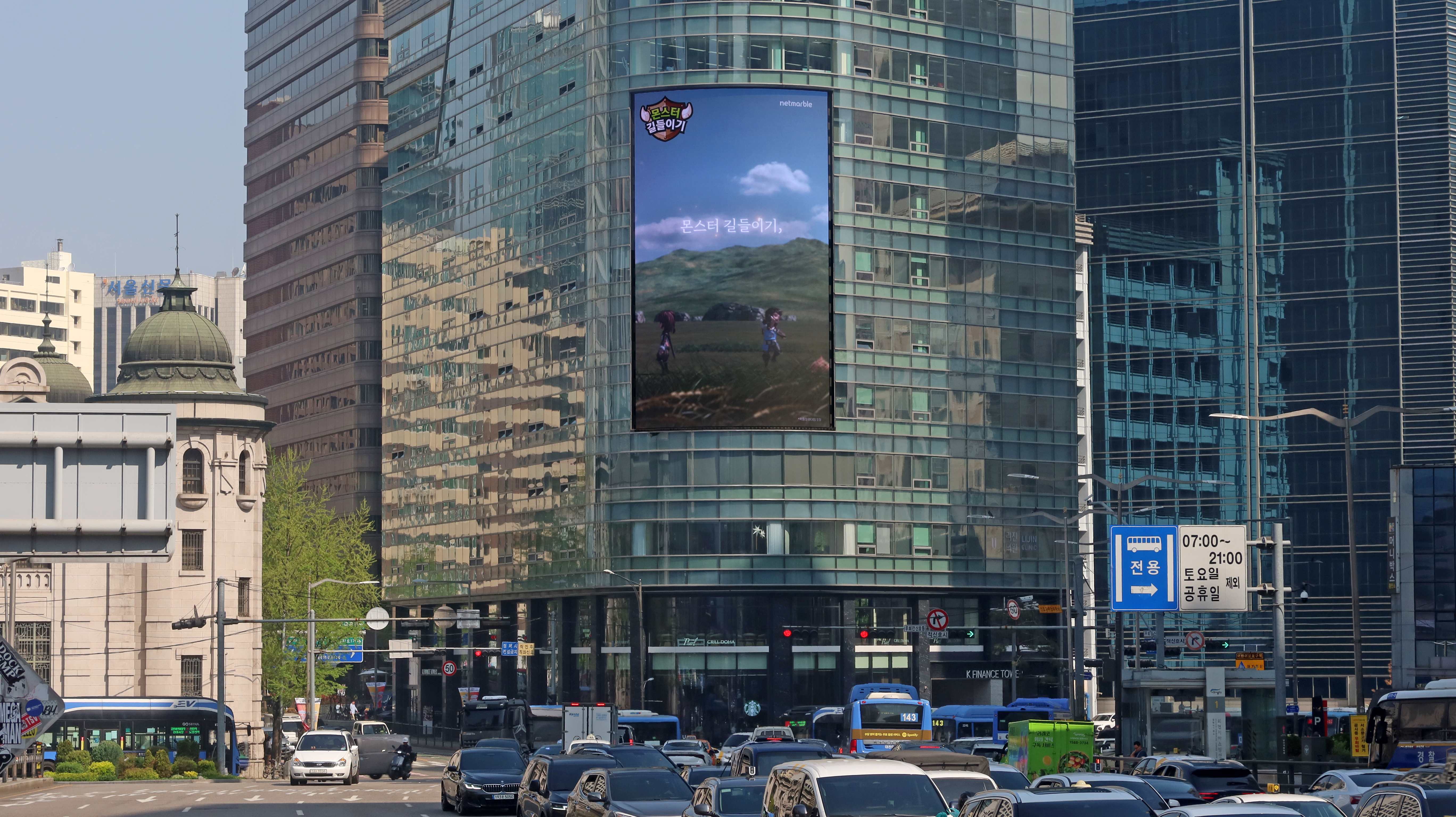 Monster Taming creative on the vertical screen of a curved glass tower above heavy traffic in central Seoul