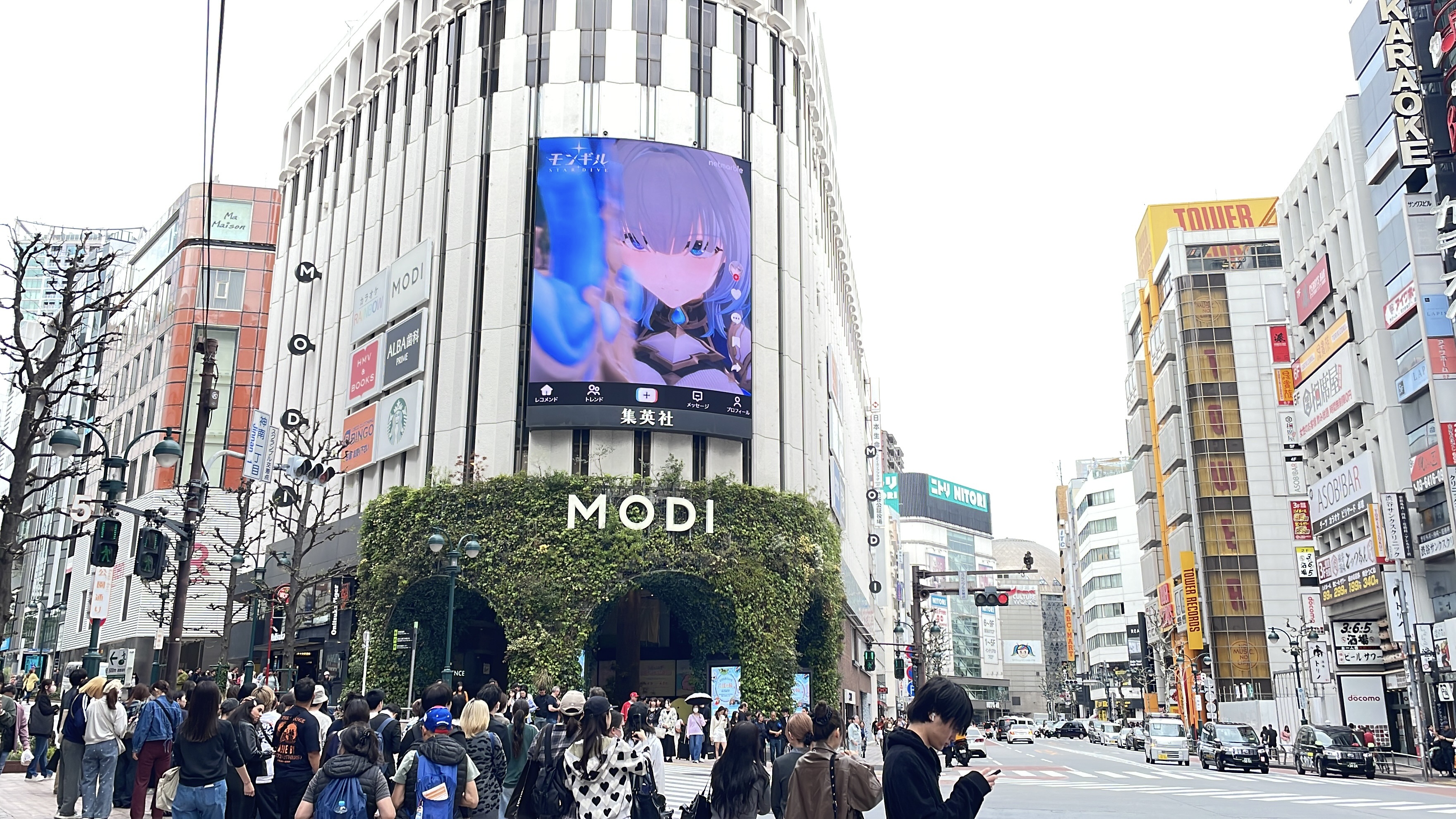 MONGIL: STAR DIVE character close-up on the curved screen of the MODI building above a crowd at the crossing, Shibuya, Tokyo