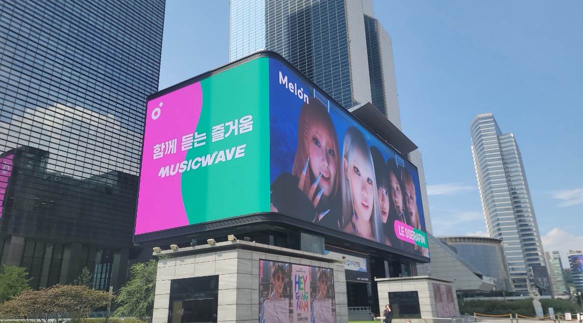Melon MUSICWAVE and LE SSERAFIM campaign on the curved COEX K-POP LIVE screen under a clear sky, Seoul