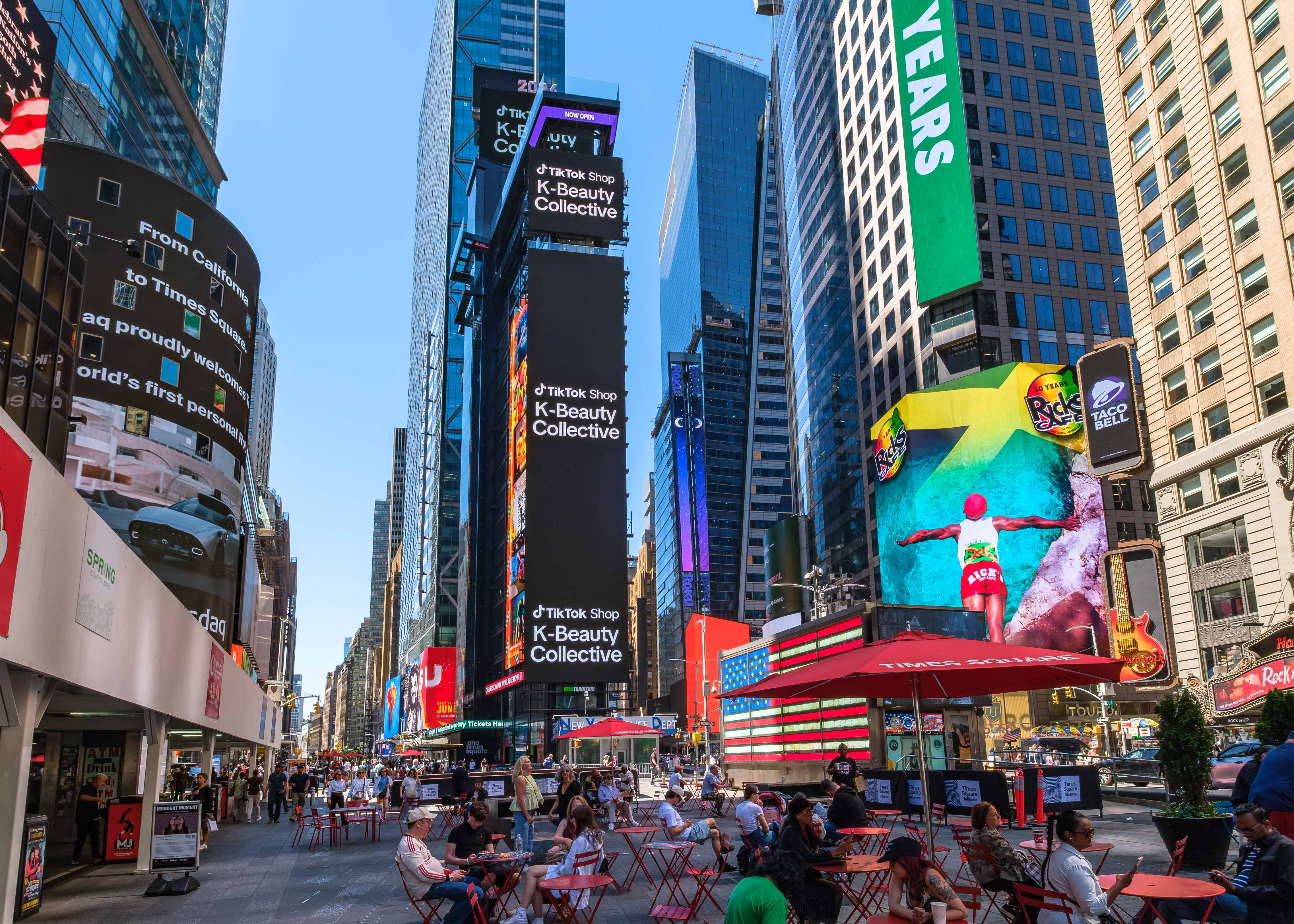 Wide daytime view of Times Square with the TikTok Shop K-Beauty Collective identity on the vertical screens above the plaza seating